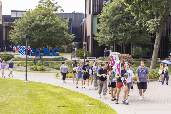 A group of Macalester first year students walks down a sidewalk behind an Orientation Leader holding a large wooden shield