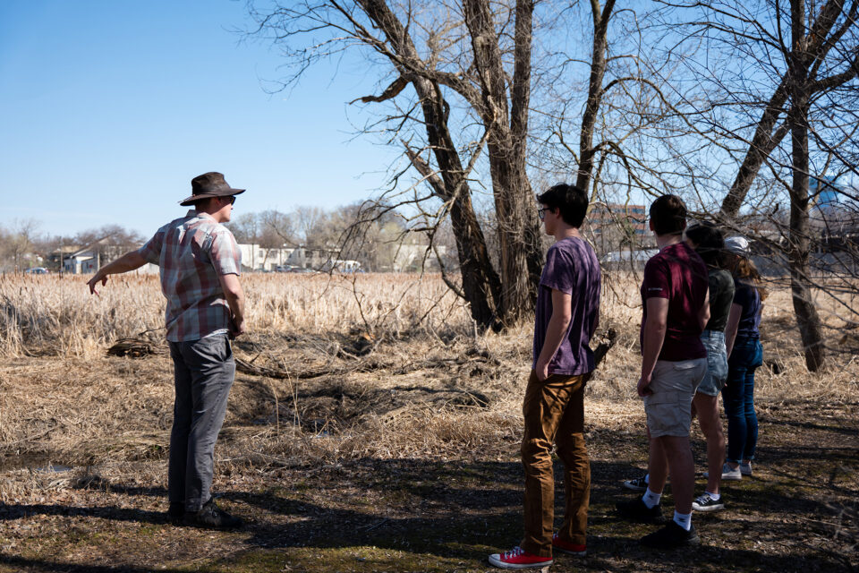 Students stand on farmland, listening to a farmer speak