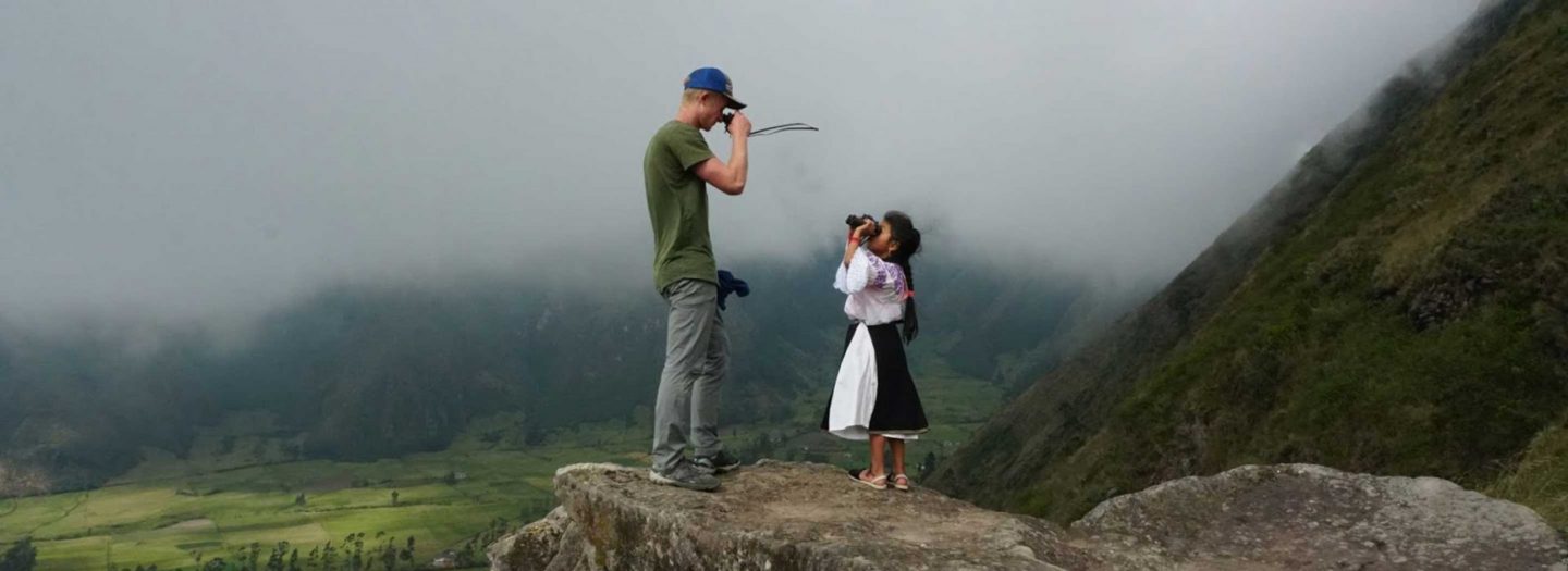 A student and a local inhabitant stare at each other with binoculars while standing atop a mountain.
