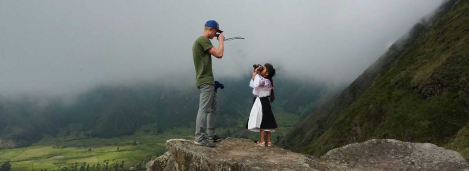 A student and a local inhabitant stare at each other with binoculars while standing atop a mountain.
