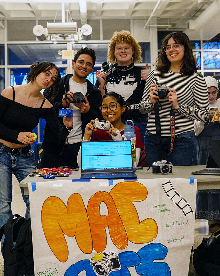 A group of mac students gather around a table at the Spring Student Org Fair, displaying film equipment.