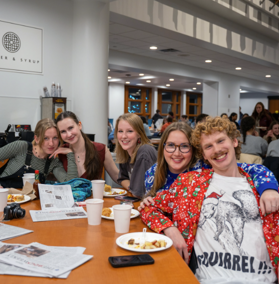 Macalester students sit around a table in Cafe Mac, smiling with their midnight breakfast spread.