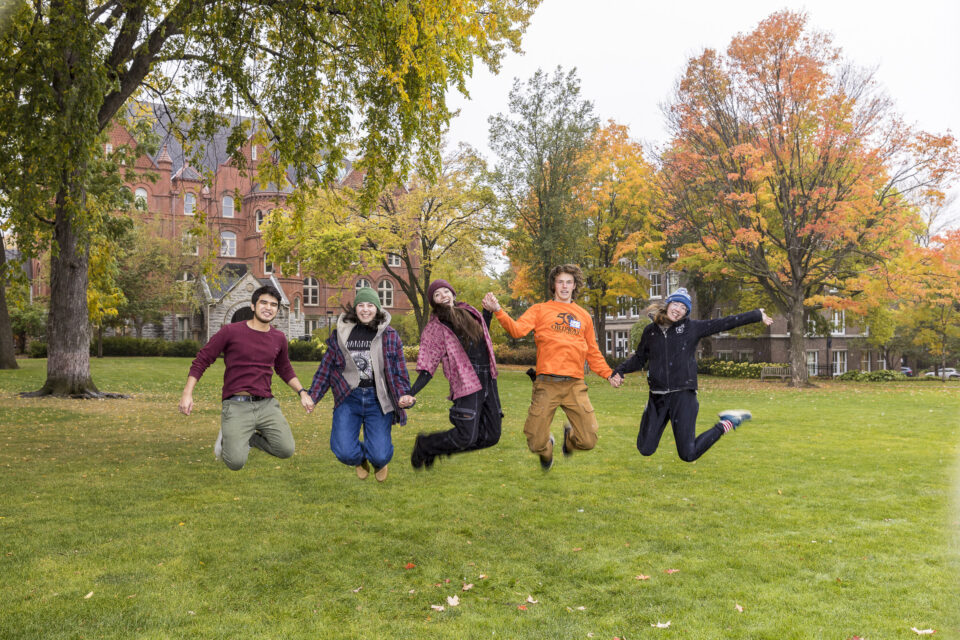 Five people jump as they pose for a photo on the Great Lawn at Macalester College. Old Main and trees with fall leaves are visible in the background.