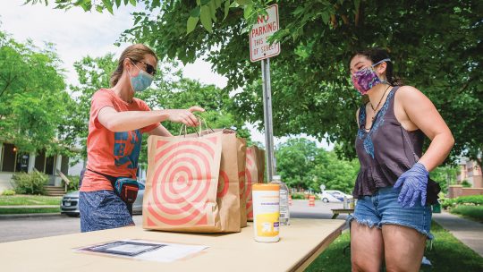 Photo of a donor placing bags of food on a table in front of a volunteer (both wearing masks)