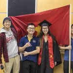 Four people smile as they stand next to the Vietnamese flag at Macalester's 2022 December Graduation.