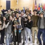 Twelve people pose for a picture with a Macalester 2022 December graduate. Many of the people are looking at the graduate and pointing to them.