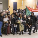 A group, including students, staff, faculty, friends, and family, poses next to the Sudanese flag at Macalester's 2022 December Graduation.