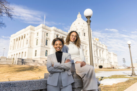 Mac students Mena Feleke and Amanda de Souza pose in front of the Minnesota State Capitol Building.