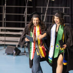 Two graduates walk with their diplomas, holding hands and smiling