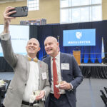 Two alumni take a selfie in front of a podium with a Macalester logo