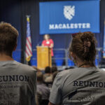 Two Reunion student workers watch a person give a speech at a podium on stage