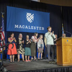 An alum stands at a podium clapping their hands while a row of people behind them clap their hands