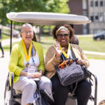 Two alumni wave from the back of a golf cart