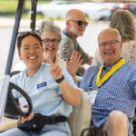 Three alumni smile and wave from a golf cart, along with a student Reunion worker who is driving
