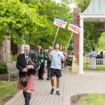 A bagpiper and a student holding signs reading 1983 and CLUSTER FUSS lead a parade