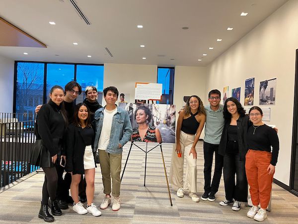 A group of nine students pose together around a large printed photo display at the Adelante photo exhibit.