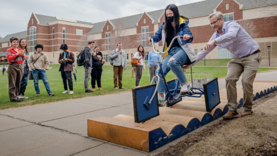 Stan Wagon helps a student ride a square-wheeled bike while other students look on