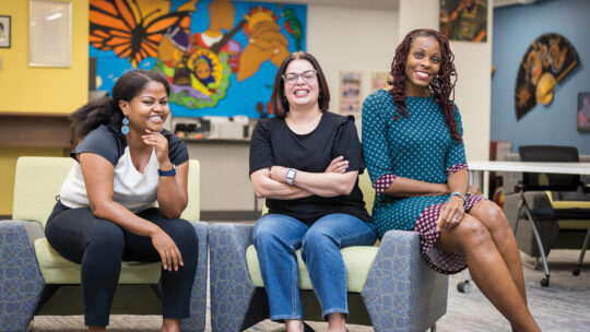 Hana Dinku, Aida Martinez-Freeman, and Karla Benson Rutten pose on chairs inside of the Lealtad-Suzuki Center for Social Justice.