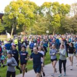 A big crowd runs the 5K race on a sidewalk and campus lawn.