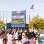 Community members gather outside and enjoy warm beverages. The scoreboard behind them is lit up with a Mac Fest photo.