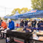Four people work the grills at a Mac Fest tailgate. Others are gathered in Macalester Athletics tents behind them.