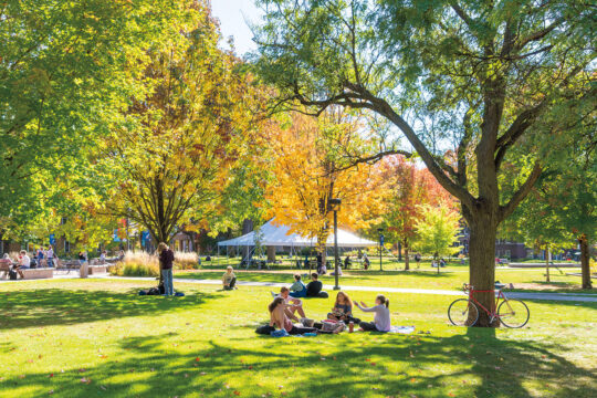 Students sit in a circle on Macalester's Great Lawn in the fall. Some trees have orange and yellow leaves. A red bike rests against a tree on the right.