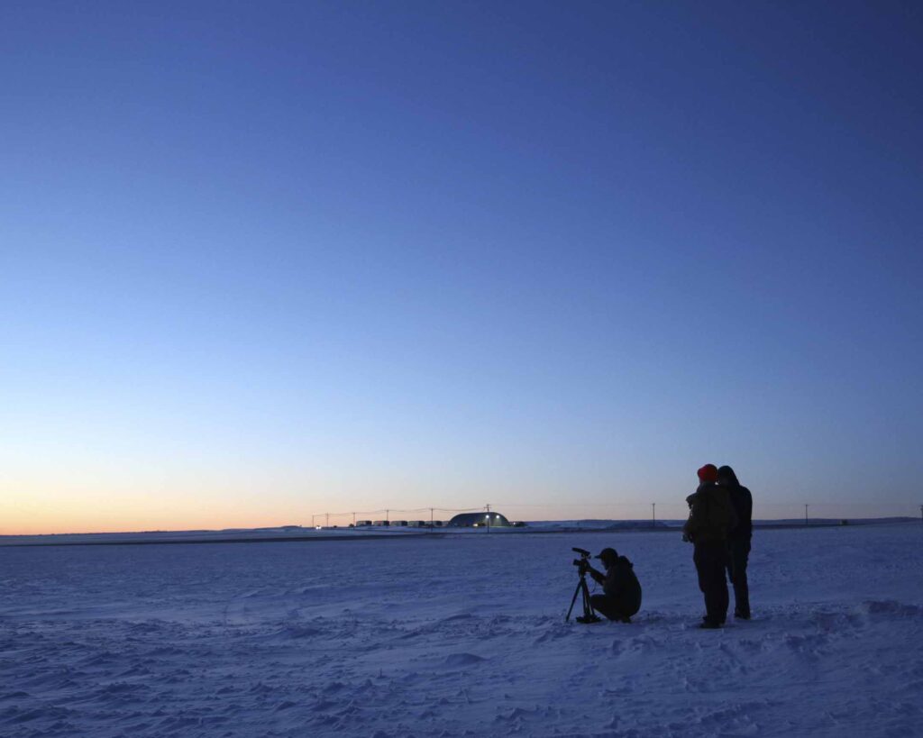 A person with a camera crouches in the snow with two people standing behind them. The final moments of the sunset are visible in the distance as well as a few small buildings and powerlines.