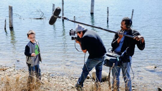 Two filmmakers, one holding a camera and one holding an overhead microphone, interview a young subject on the shore next to a body of water.
