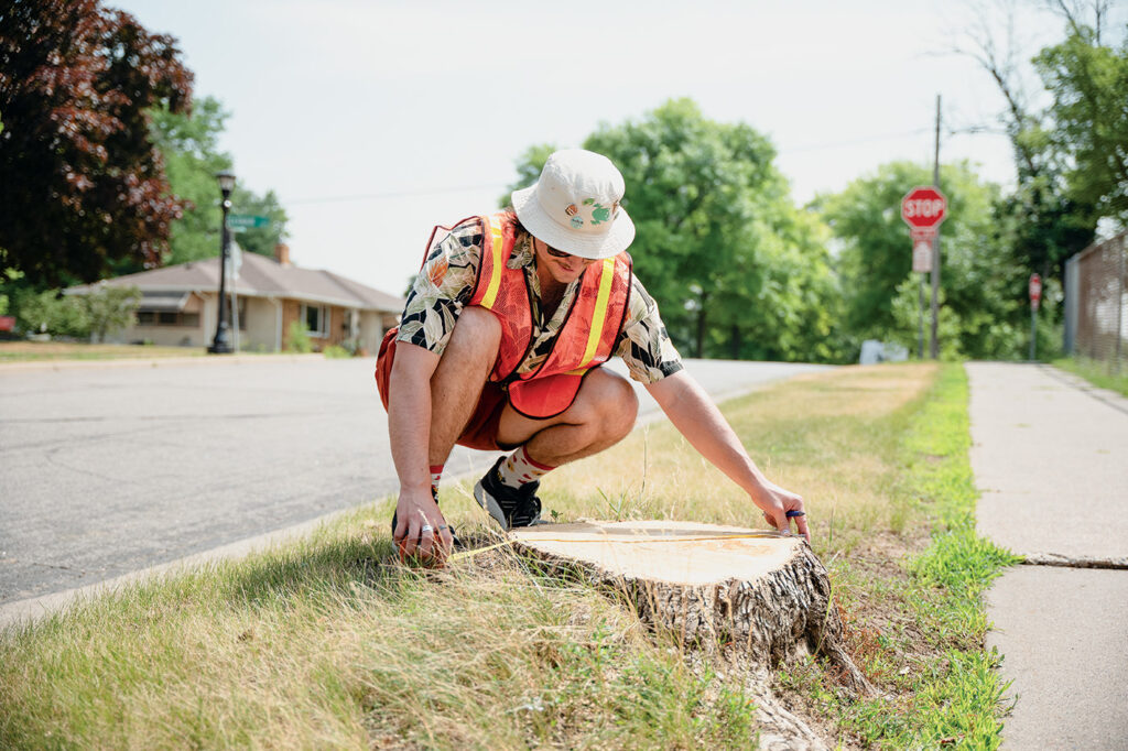 A person wearing an orange vest measures a tree stump next to a residential street.