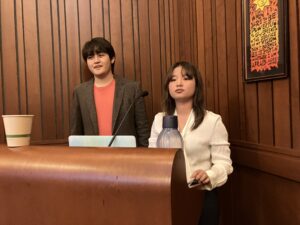 Two students, one male and one female, stand in front of a podium with a microphone. Both are in business casual outfits. The male student on the left is smiling or in mid-speech, and the female student on the right appears to be listening to a question from the audience (not pictured).