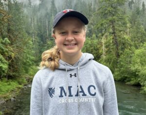 Erin Franke '23 stands in front of a small stream surrounded by trees in a gray Macalester cross country sweatshirt and navy Twins baseball cap. 