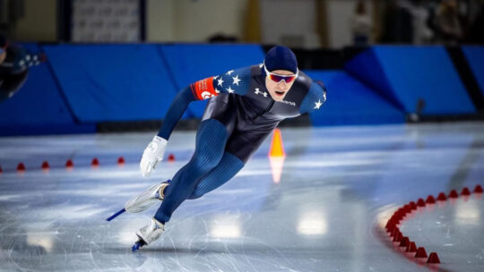 A man on skates rounds the corner of an ice rink in a slick blue uniform, with white stars on the shoulders and a red arm band. His head is covered except for his face. He wears reflective glasses.