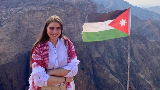 Chloe Vasquez poses in front of a scenic canyon beside the Jordanian national flag.