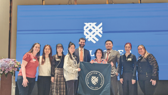 A group of Macalester alumni and staff pose together in Beijing, holding a Macalester College banner in front of a large Macalester logo screen.