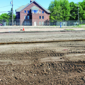 Exposed streetcar railway tracks beneath Grand Avenue asphalt during resurfacing project, with a brick building in the background.