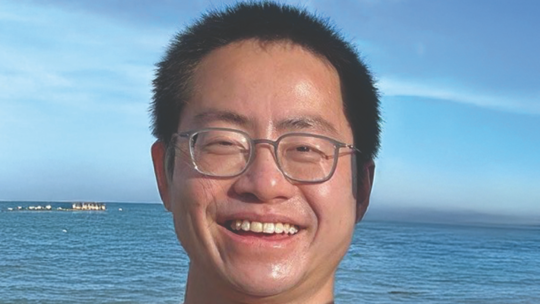 Close-up of Yulun Li ’14 smiling outdoors with the ocean and blue sky in the background.