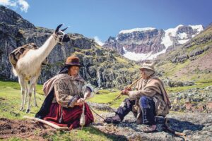 Two women sit in a mountain valley, with an alpaca standing nearby, in this scene from Wiñaypacha (Eternity), a 2018 Peruvian drama directed by Óscar Catacora.