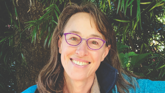 Smiling Jennifer Wofford with shoulder-length brown hair wearing purple glasses and a blue jacket, standing outdoors in front of a tree and green foliage.