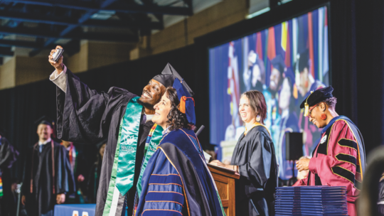 Graduate in cap and gown takes a selfie with Macalester College President Suzanne M. Rivera on stage during the 2025 Commencement ceremony, with faculty and diplomas visible in the background.