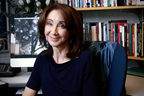 Professor Ruthanne Kurth-Schai sits at her desk, smiling warmly in front of a bookshelf and computer, reflecting her lifelong dedication to education and civic learning.