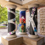 Left to right: a black pillar with white curved stripes, a pillar with a Native figure wearing orange and brown and dancing against a sunset, a black pillar with a white X shape and purple, yellow, and blue symbols in the middle of the X