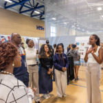 A student presents their academic poster to a group of visitors during the Mac Fest Academic Summer Showcase inside the Leonard Center gym.