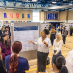 A student speaks to a crowd gathered around their research poster at the Academic Summer Showcase during Mac Fest 2025.