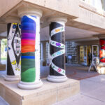 Left to right: a black pillar with a white X shape and purple, yellow, and blue symbols in the middle of the X, a pillar painted to resemble a sunset, a black pillar with white curved stripes, the door to DeWitt Wallace Library