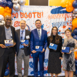 Award recipients pose with their plaques in front of a festive orange and blue Macalester backdrop during Mac Fest 2025