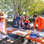 Volunteers in orange Macalester shirts serve food from the grill to guests at the M Club Cookout during Mac Fest 2025.