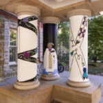 Left to right: a white pillar with black curved stripes, a black pillar with a Native figure wearing white, a white pillar with a white X shape and purple, yellow, and blue symbols in the middle of the X