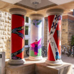 Left to right: a red pillar with black curved stripes, a white pillar with a Native figure dancing and wearing a purple and pink outfit, a red pillar with a white X shape and purple, yellow, and blue symbols in the middle of the X