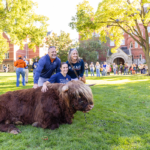 A group poses next to Hartnett the mini Highland Coo lying on the grass during Mac Fest 2025.