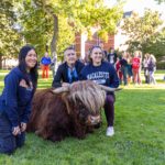 Three attendees kneel and smile beside Hartnett the mini Highland Coo during Mac Fest 2025.